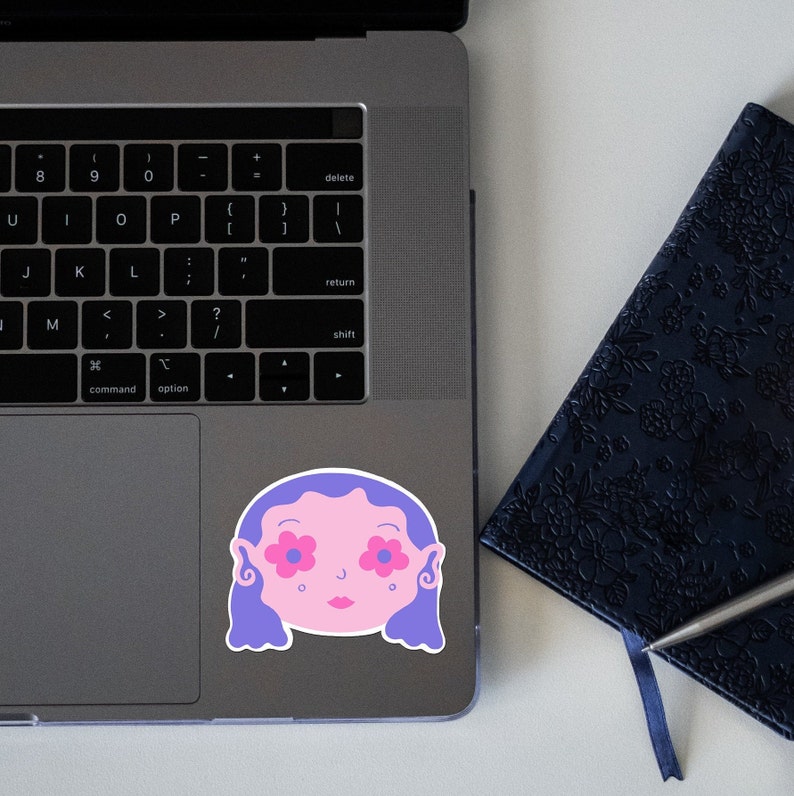May include: A sticker of a cartoon face with purple hair and pink flowers for eyes. The sticker is on a silver laptop.