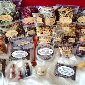 Peut inclure: Une collection d'herbes et de fleurs séchées dans des sachets en plastique transparent, étiquetés avec leurs noms, tels que Bardane, Bourgeon de rose et Sauge blanche. Il y a aussi de petits pots de divers ingrédients, dont du charbon de bois, du sel de l'Himalaya et du sel noir. Les articles sont disposés sur une surface moelleuse blanche.
