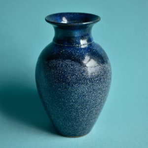 May include: A small, speckled blue ceramic vase with a flared rim. The vase has a dark blue top that fades into a lighter blue with white speckles. The vase is set against a light blue background.