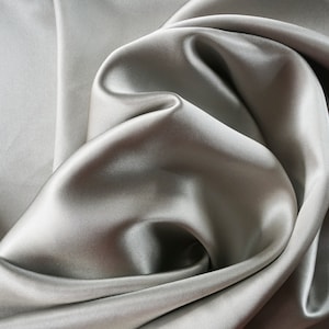 Può includere: Un tessuto liscio color argento drappeggiato in morbide pieghe. Il tessuto ha un aspetto setoso e una sensazione lussuosa.