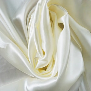 Könnte beinhalten: Nahaufnahme eines glatten, elfenbeinfarbenen Satin-Stoffes, der in weichen Falten drapiert ist.