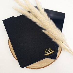 Könnte beinhalten: Schwarzes Ledernotizbuch mit goldenen Buchstaben, die "GIA" mit einem goldenen Blattdesign lesen. Das Notizbuch liegt auf einer Holzoberfläche mit einem Tablet und einem Zweig getrockneten Pampasgrases.