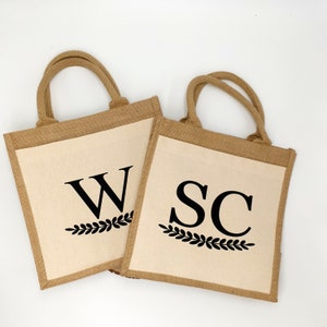 Könnte beinhalten: Zwei braune Jute-Tragetaschen mit weißem Stoff-Innenfutter. Die Taschen haben schwarze Beschriftung mit einem Blattdesign. Eine Tasche sagt "W" und die andere sagt "SC".