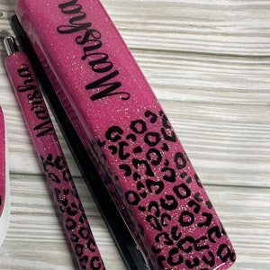 May include: A pink glitter stapler, pen, and pencil holder set. The stapler and pen are decorated with black leopard print spots. The name "Marsha" is written in black script on the stapler and pen.