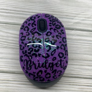 May include: Purple wireless computer mouse with a black leopard print design and the name "Bridget" in cursive. The mouse has a black scroll wheel and base, and is set against a light wood grain background.