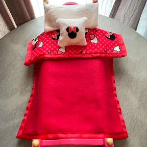 May include: A miniature red bed with a matching blanket and pillows. The blanket has a red polka dot pattern with Minnie Mouse images. The small pillow has a Minnie Mouse design. The bed frame is pink.