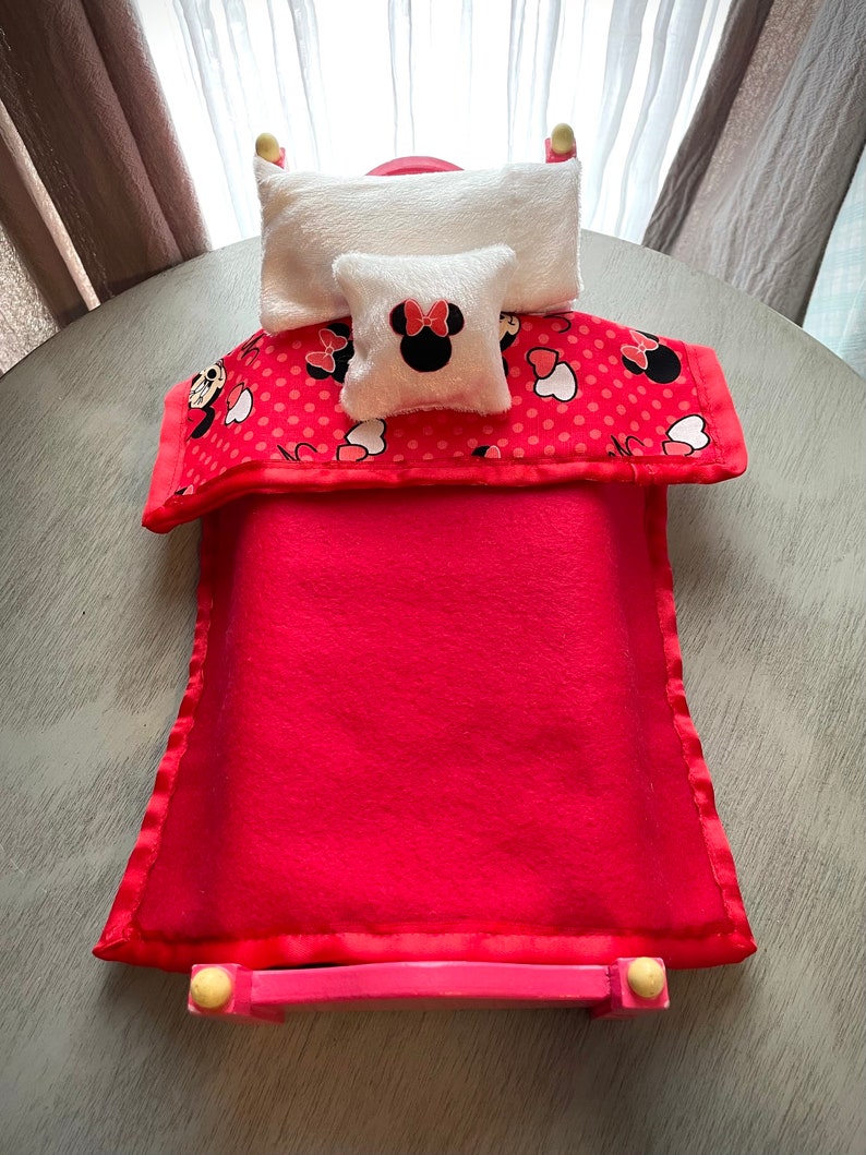 May include: A miniature red bed with a matching blanket and pillows. The blanket has a red polka dot pattern with Minnie Mouse images. The small pillow has a Minnie Mouse design. The bed frame is pink.