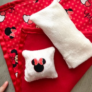 May include: A red blanket with a satin edge, featuring a Minnie Mouse pattern with polka dots and hearts. Two white plush items, a rectangular pillow and a square pillow with a Minnie Mouse face, are on top of the blanket.