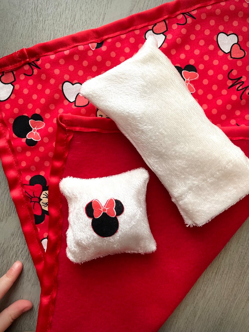 May include: A red blanket with a satin edge, featuring a Minnie Mouse pattern with polka dots and hearts. Two white plush items, a rectangular pillow and a square pillow with a Minnie Mouse face, are on top of the blanket.