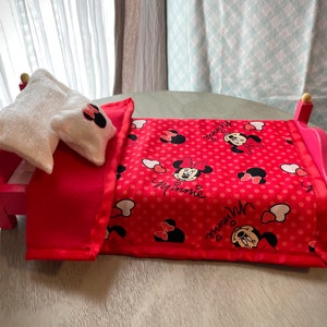 May include: A miniature pink wooden bed with a red blanket and two white pillows. The blanket features a pattern of Minnie Mouse and polka dots. The pillows have Minnie Mouse bows. The bed frame has rounded posts with yellow accents.