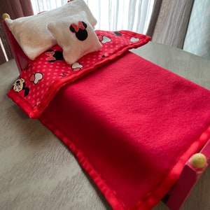 May include: A small red bed with a red blanket, a patterned comforter, and two white pillows. One pillow features a Minnie Mouse design. The bed frame is pink with yellow accents on the posts. The comforter has a red background with white polka dots and Minnie Mouse graphics.