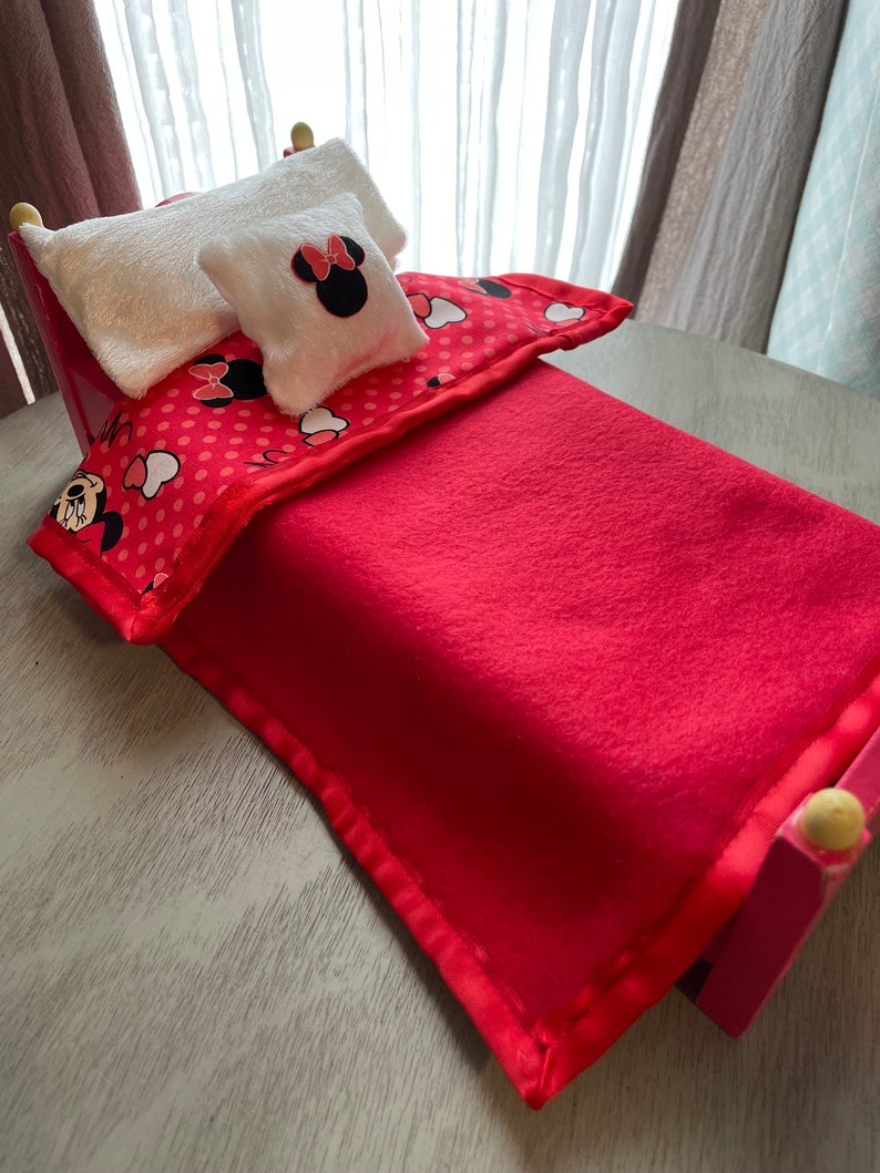 May include: A small red bed with a red blanket, a patterned comforter, and two white pillows. One pillow features a Minnie Mouse design. The bed frame is pink with yellow accents on the posts. The comforter has a red background with white polka dots and Minnie Mouse graphics.