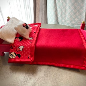 May include: A small pink bed with a red blanket and a pillow. The blanket features a red polka dot pattern with Minnie Mouse graphics. The pillow is white with a Minnie Mouse bow. The bed frame is pink with gold detailing.