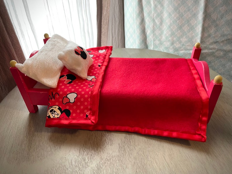 May include: A small pink bed with a red blanket and a pillow. The blanket features a red polka dot pattern with Minnie Mouse graphics. The pillow is white with a Minnie Mouse bow. The bed frame is pink with gold detailing.