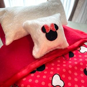 May include: A miniature bed set with a red blanket, a white pillow, and a smaller pillow featuring a Minnie Mouse design. The bed set includes a red blanket with a polka dot pattern and a white pillow with a Minnie Mouse design.