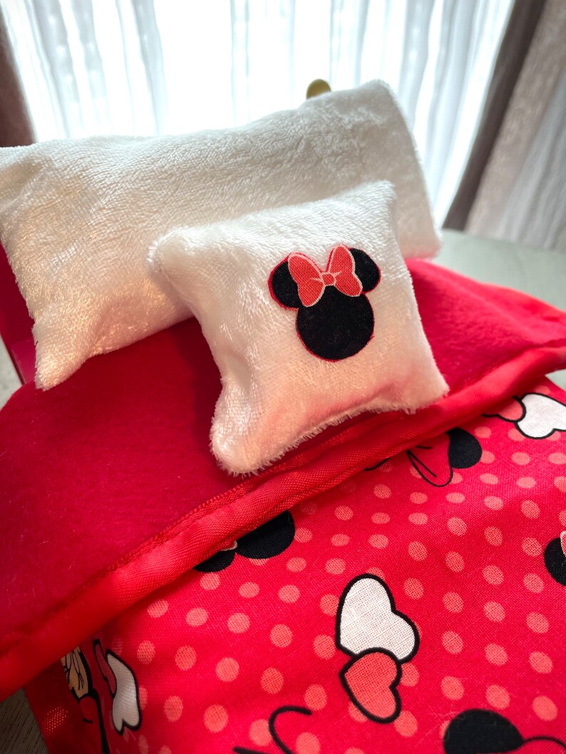 May include: A miniature bed set with a red blanket, a white pillow, and a smaller pillow featuring a Minnie Mouse design. The bed set includes a red blanket with a polka dot pattern and a white pillow with a Minnie Mouse design.