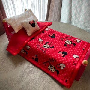 May include: A miniature pink bed with a red blanket featuring a Minnie Mouse pattern. Two white pillows, one rectangular and one square, complete the bedding set. The bed frame is pink with rounded wooden accents.
