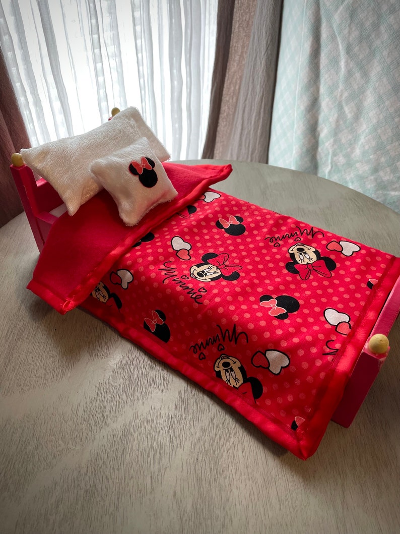 May include: A miniature pink bed with a red blanket featuring a Minnie Mouse pattern. Two white pillows, one rectangular and one square, complete the bedding set. The bed frame is pink with rounded wooden accents.