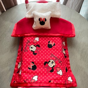 May include: A small pink bed with a red headboard and footboard. It features a white pillow and a small pillow with a Minnie Mouse design. The blanket is red with white polka dots and Minnie Mouse graphics.