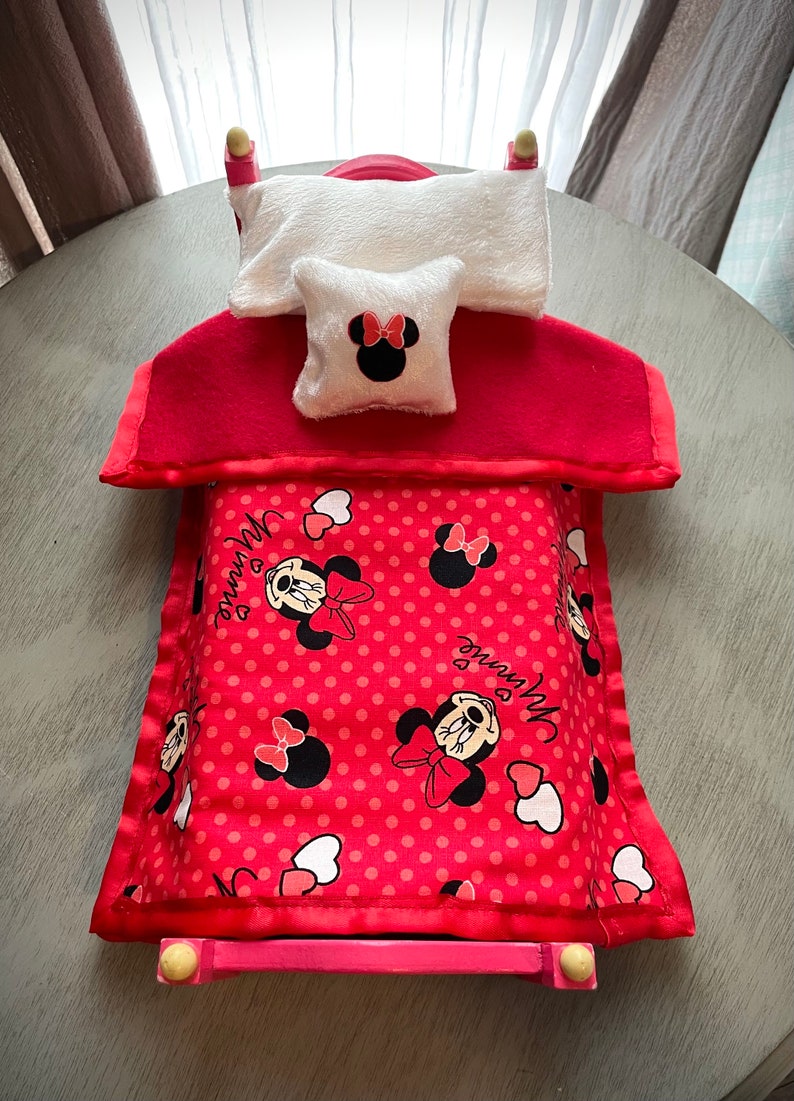 May include: A small pink bed with a red headboard and footboard. It features a white pillow and a small pillow with a Minnie Mouse design. The blanket is red with white polka dots and Minnie Mouse graphics.