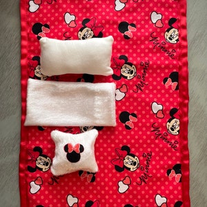 May include: A red and white Minnie Mouse themed baby blanket with three white plush items. The blanket has a red satin border and is patterned with Minnie Mouse faces, bows, and hearts. The word "Minnie" is written in cursive.