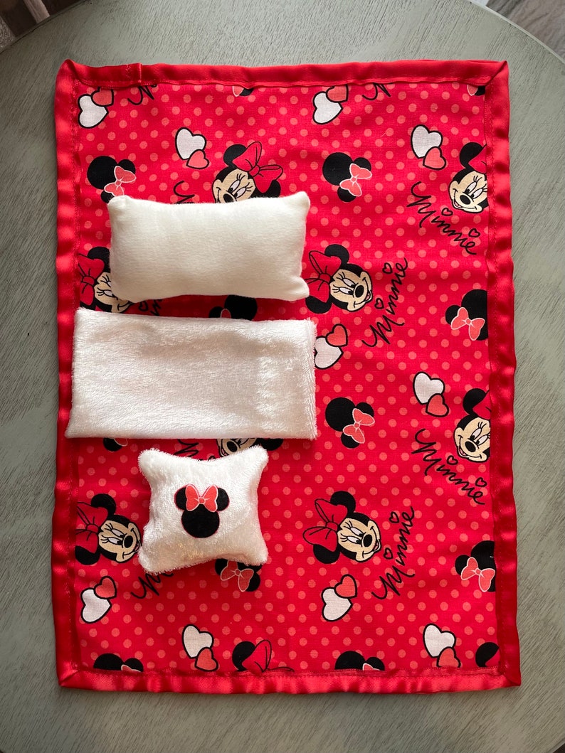 May include: A red and white Minnie Mouse themed baby blanket with three white plush items. The blanket has a red satin border and is patterned with Minnie Mouse faces, bows, and hearts. The word "Minnie" is written in cursive.