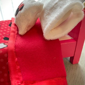 May include: A miniature pink bed with a red blanket and a pillow. The blanket has a satin edge and the pillow has a black and red design. A white plush sock is on the bed.