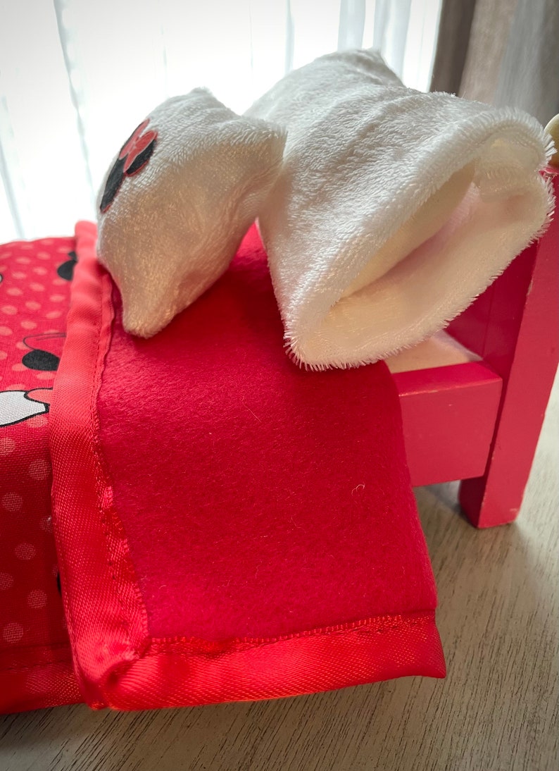 May include: A miniature pink bed with a red blanket and a pillow. The blanket has a satin edge and the pillow has a black and red design. A white plush sock is on the bed.