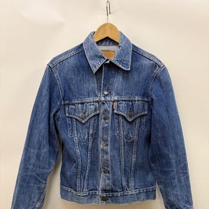 Op de afbeelding: Een blauw spijkerjack met een klassiek ontwerp, met een kraag, knoopsluiting en twee borstzakken. De jas hangt aan een houten hanger. De jas heeft oranje stiksels en oranje labels.