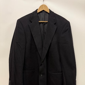 May include: A black blazer hanging on a wooden hanger. The blazer has a two-button closure, notched lapels, and two front pockets. The sleeves have three buttons at the cuffs. The blazer is made of a dark, possibly wool, fabric.