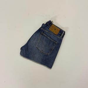 May include: Folded blue denim jeans with a faded wash. The jeans feature a classic five-pocket design and a leather patch on the back waistband. The jeans are neatly folded, displaying the back pockets and the brand label.