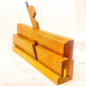 Könnte beinhalten: Ein hölzerner Hobel mit dunkelbrauner Oberfläche. Der Hobel hat eine Klinge aus Metall und einen Griff aus Holz. Der Hobel besteht aus mehreren zusammengefügten Holzstücken.