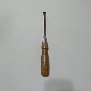 May include: A small, wooden-handled chisel with a metal blade. The handle is made of light brown wood with a dark brown grain pattern. The blade is silver and has a sharp point.