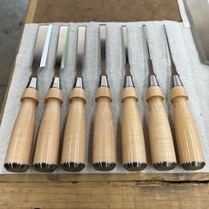May include: A set of eight wood carving chisels with wooden handles. The chisels are arranged in a row on a white surface.