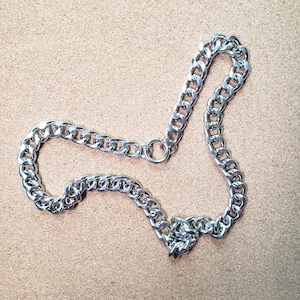 May include: A silver-colored chain necklace with large, interlocking links. The necklace is laid out on a light brown surface. The chain has a circular clasp and a lobster claw clasp.