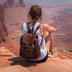 Puede incluir: Una persona con el pelo largo y castaño, que lleva un top azul claro y una mochila de cuero marrón, se sienta en un acantilado rocoso con vistas a un vasto paisaje desértico.