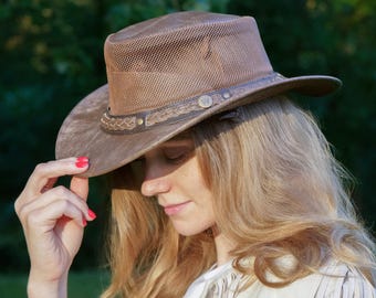 May include: A brown hat with a mesh crown and a wide brim. The hat features a decorative band around the base of the crown and a small metal emblem. The hat is a neutral color and appears to be made of a durable material.
