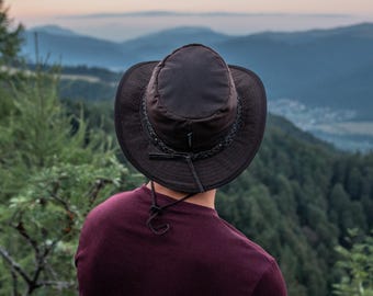 May include: A dark brown hat with a wide brim and a braided band around the crown. The hat has a chin strap and is worn by a person looking out at a mountain range. The person is wearing a maroon shirt.