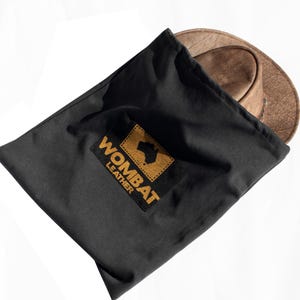 May include: A black fabric bag with a brown leather hat inside. The bag has a yellow logo that says "WOMBAT LEATHER" with a silhouette of Australia inside the logo.