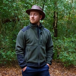May include: A man wearing a green fleece jacket with black accents, a brown hat, and blue jeans. He is standing in a forest.