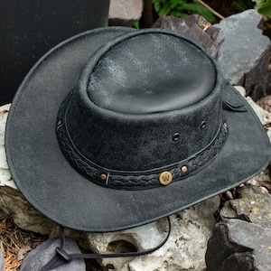 Puede incluir: Un sombrero vaquero de cuero negro con una banda trenzada y una hebilla de metal con la letra "W". El sombrero tiene un aspecto desgastado y está sobre una pila de piedras.