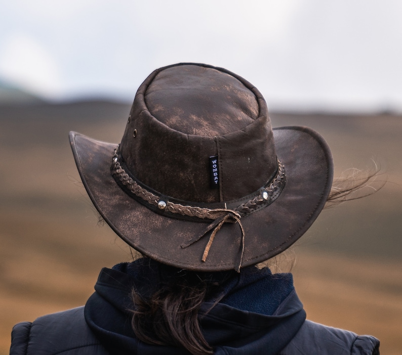 May include: A brown leather cowboy hat with a braided band and a worn look. The hat has a label that says "WOMBAT" on the side.
