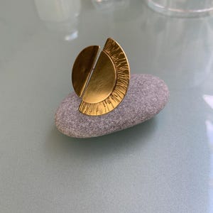 May include: A gold ring with a geometric design. The ring is made of two half circles that are joined together. The half circle on the right has a textured surface.