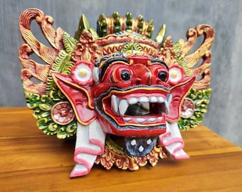 Può includere: Una maschera balinese rossa, verde e dorata con intagli intricati e un'espressione feroce. La maschera ha denti grandi e bianchi e una lingua nera.