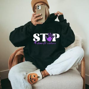 Könnte beinhalten: Schwarzer Kapuzenpullover mit dem Wort "STOP" in weißen Blockbuchstaben, einem lila Handabdruck und einem lila Band. Die Worte "domestic violence" stehen unter dem Wort "STOP". Der Kapuzenpullover wird von einer Person getragen.