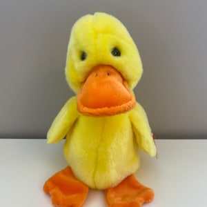 May include: A yellow plush duck with an orange beak and feet. The duck has black eyes and a soft, fuzzy texture.