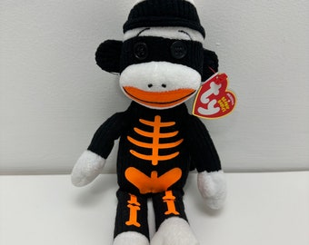 Puede incluir: Un mono de peluche negro y blanco con un diseño de esqueleto naranja lleva un gorro a rayas negro y blanco con un pompón naranja. El mono está sentado con las piernas cruzadas y tiene una cara blanca con ojos negros de botón. El mono tiene una etiqueta de Ty Beanie Babies unida a su brazo.