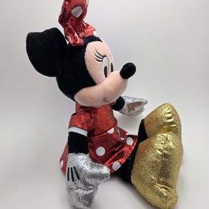 May include: A plush Minnie Mouse doll in a seated position. The doll has a red dress with white polka dots, a matching bow, and gold glitter shoes. Her gloves are silver glitter, and her ears are black.