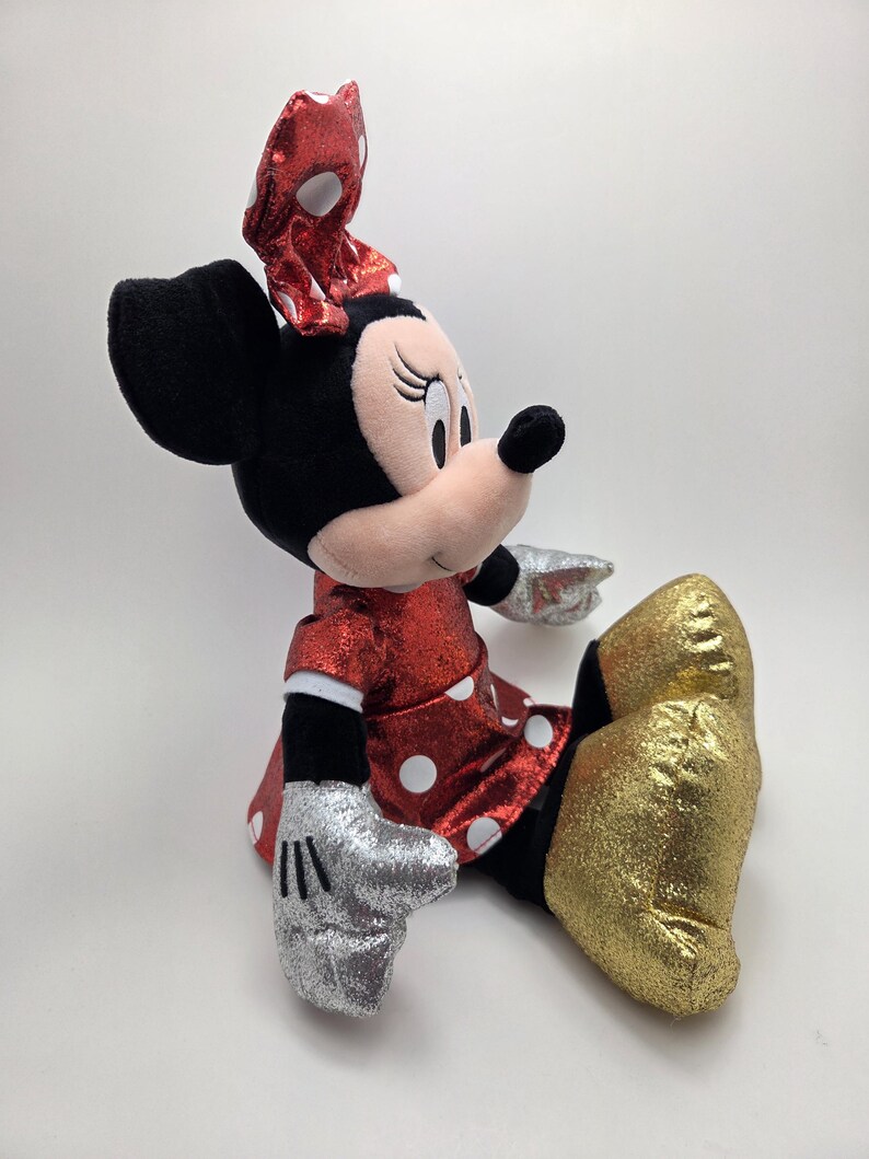 May include: A plush Minnie Mouse doll in a seated position. The doll has a red dress with white polka dots, a matching bow, and gold glitter shoes. Her gloves are silver glitter, and her ears are black.