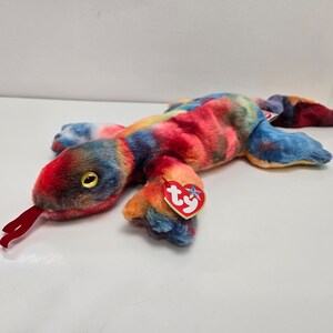 May include: A colorful plush toy lizard with a tie-dye pattern in shades of red, orange, blue, and yellow. The lizard has a red tongue, yellow eyes, and a red heart-shaped tag.